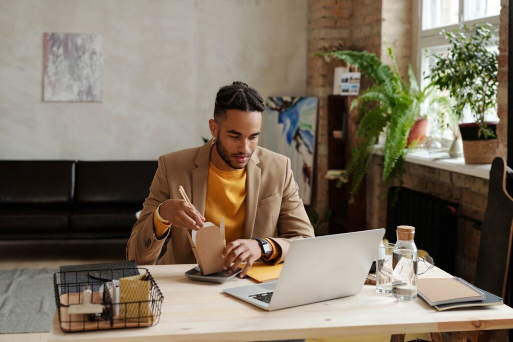 A man enjoys lunch at his workspace while using a laptop in a modern office setting.