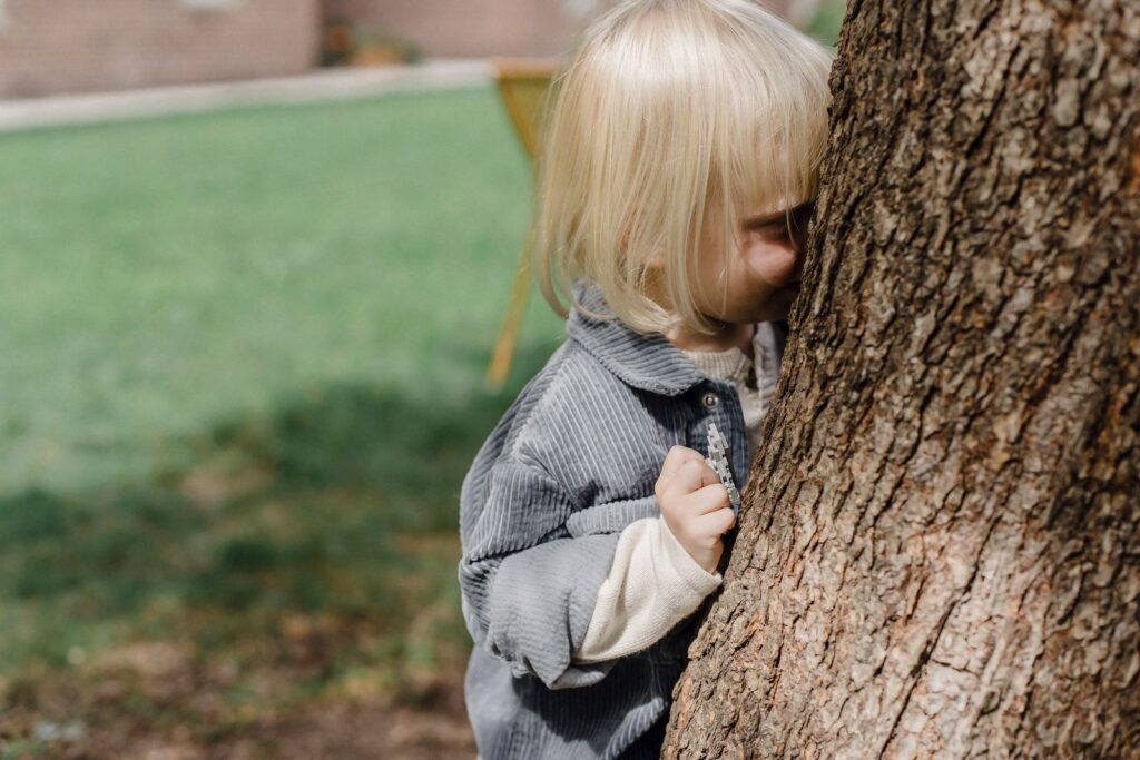 Side view of adorable little kid with blond hair hiding behind tree while playing hide and seek in park on blurred background