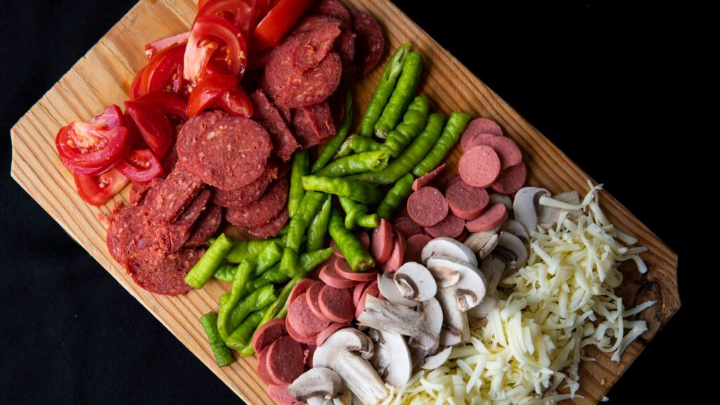 From above of meat sausages and vegetables with grated cheese placed on wooden cutting board on black background