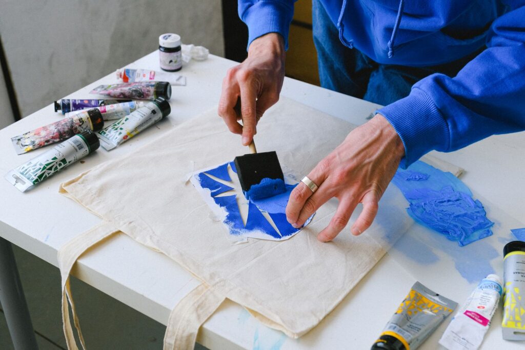 From above of unrecognizable male designer using brush and stencil to transfer sketch with blue paint on textile bag while standing at table with tubes of paints