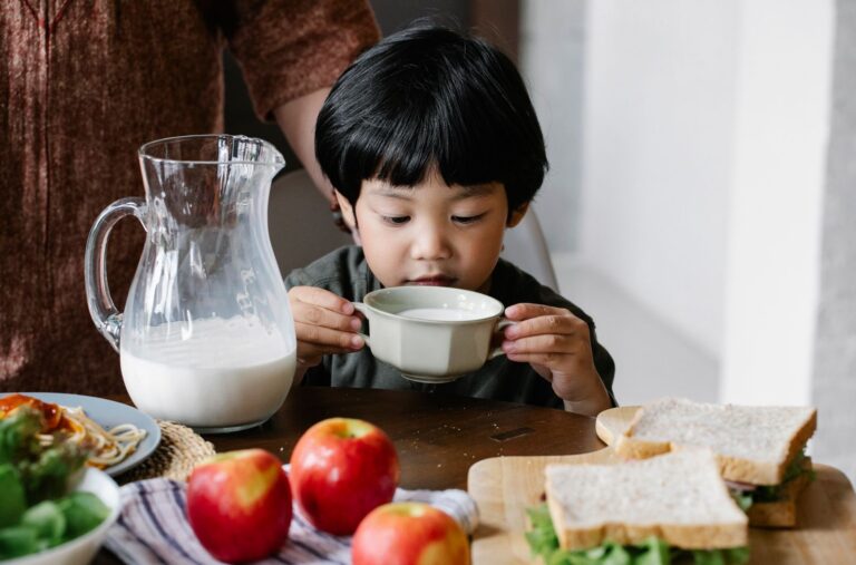 Asian little boy drinking milk from mug while sitting at served table in kitchen with crop anonymous woman behind back