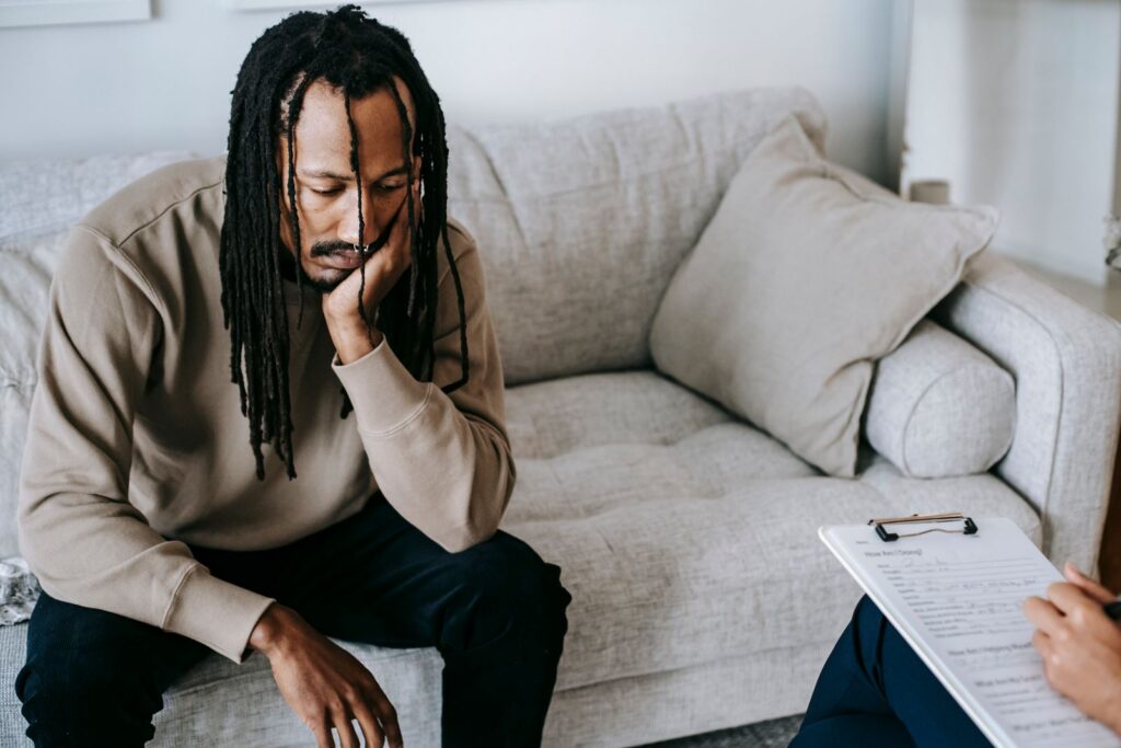 Upset African American male with dreadlocks leaning on hand and looking down while sitting on couch near crop psychologist with clipboard