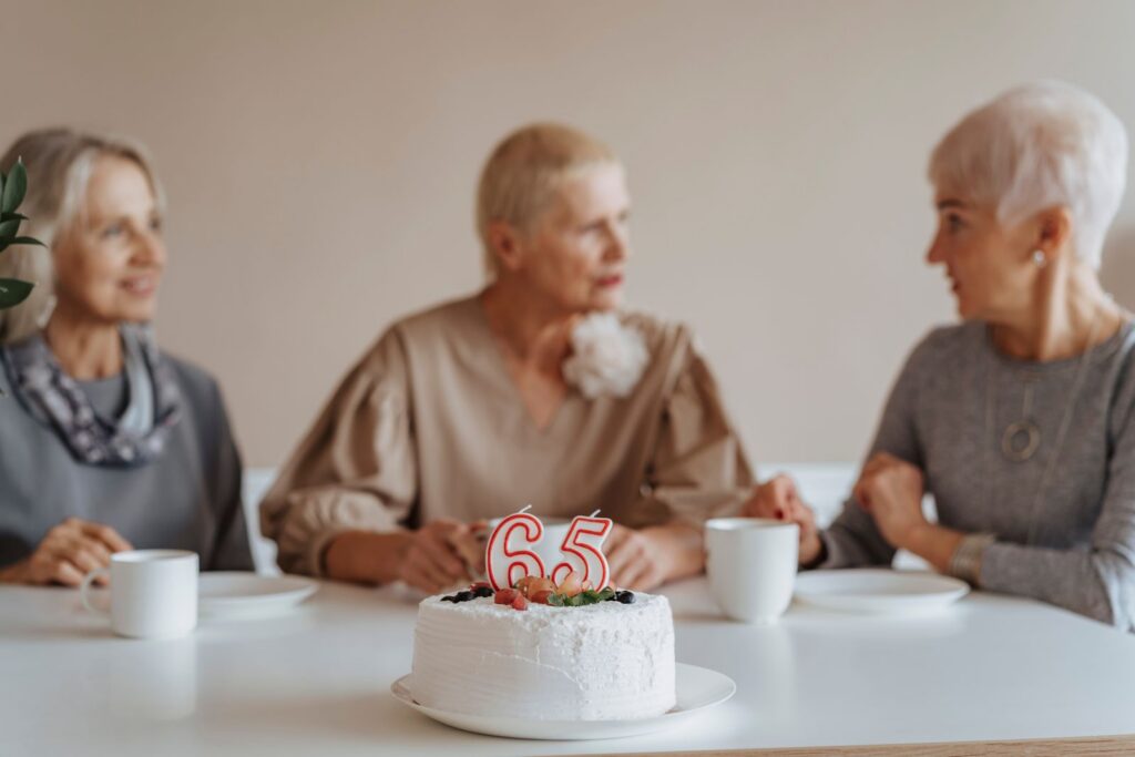 Three senior women celebrate a 65th birthday with cake and coffee indoors.