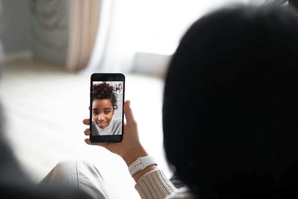 Back view of anonymous couple communicating with African American kid via video chat on modern smartphone while sitting in living room on blurred background