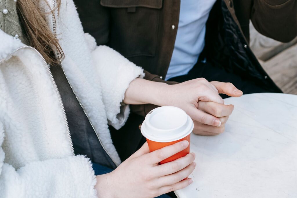 From above of crop faceless lady wearing warm jacket holding hands with boyfriend while drinking coffee to go in street cafe in daytime