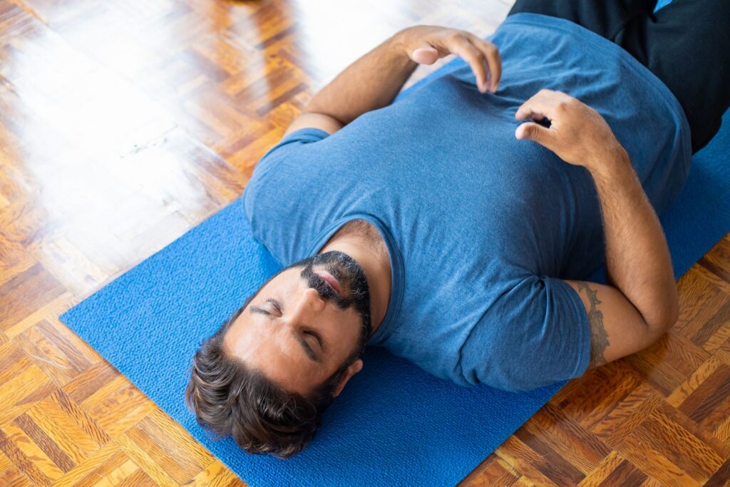 Adult man lying on a blue yoga mat indoors with eyes closed, focusing on relaxation and mindfulness.