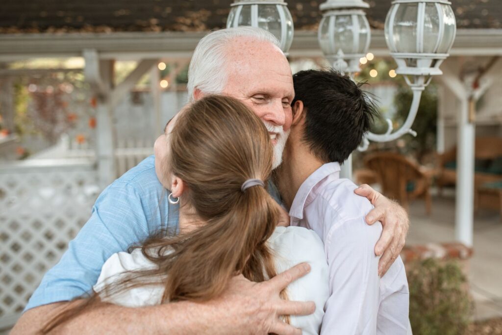 An elderly man joyfully hugs two younger adults in a cozy outdoor setting filled with affection.