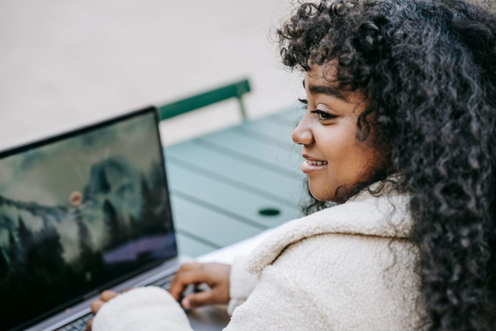 Back view content young African American female in white coat using modern netbook while sitting at table in city park and looking away with smile