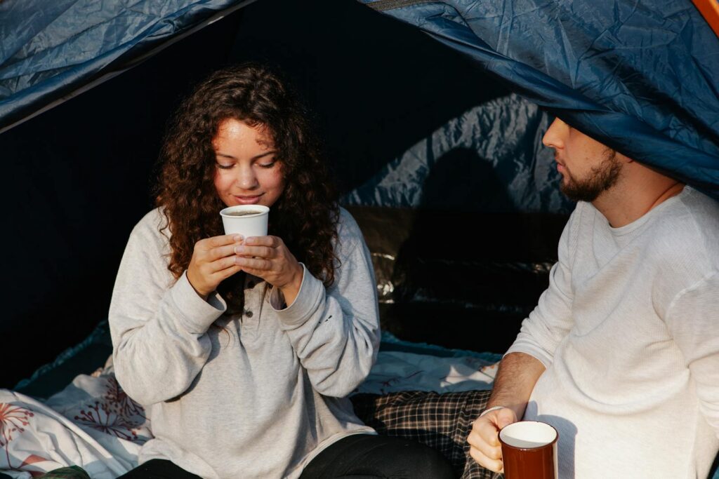 Young gentle female tourist enjoying hot drink near crop male partner while spending time in campsite