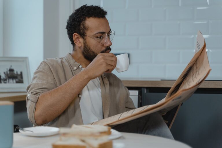 Bearded man with glasses reads newspaper while sipping coffee in a modern kitchen.