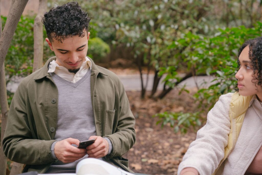 Hispanic happy boyfriend surfing internet on mobile phone while focused crop girlfriend watching in park with green plants
