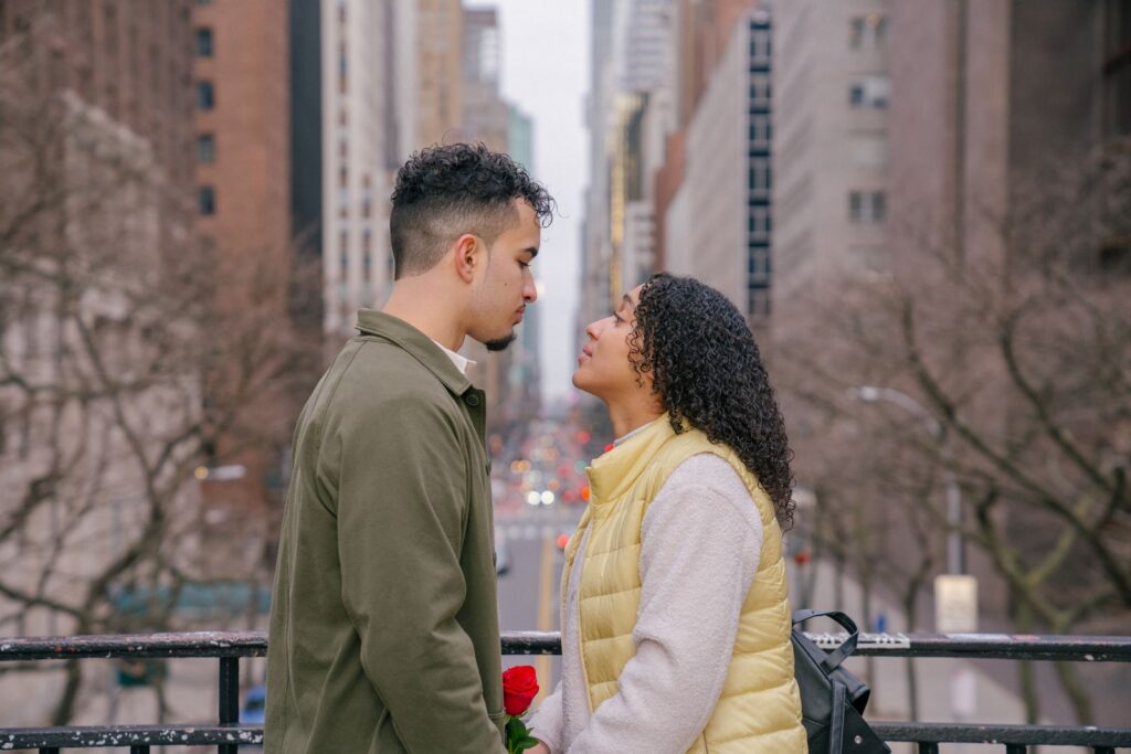 Side view of romantic Hispanic couple in casual clothes looking at each other while standing on bridge near metal fence against city street in daytime