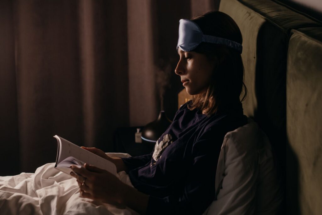 Woman wearing a sleep mask reads a book in bed at night, creating a calm and restful ambiance.