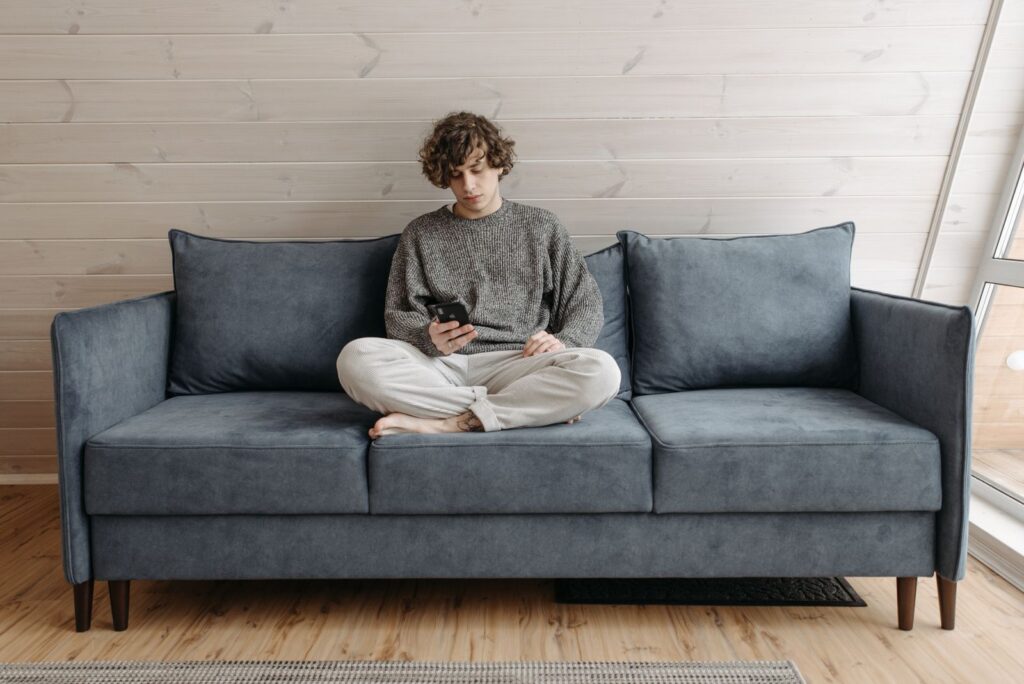 Casual young man sitting on a blue sofa using a smartphone in a cozy indoor setting.