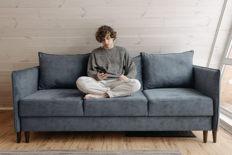 Casual young man sitting on a blue sofa using a smartphone in a cozy indoor setting.
