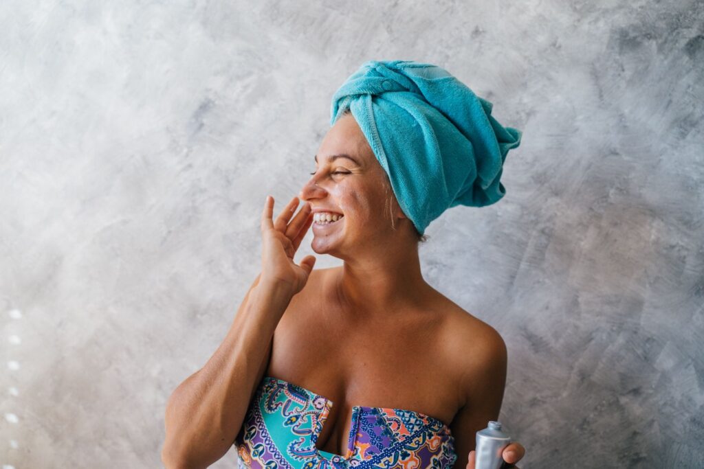 Happy woman with towel on head applying facial cream, embracing skincare routine indoors.