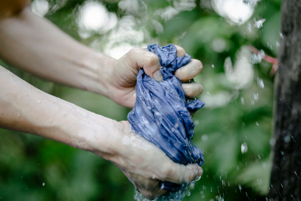 Hands of faceless person squeezing wet blue fabric under water drops while standing on street near green plants on blurred background