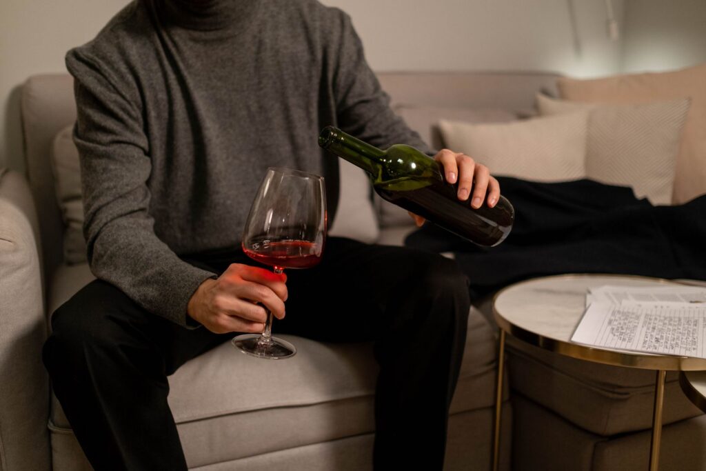 Person pouring red wine into a glass while sitting on a couch, conveying relaxation and comfort.