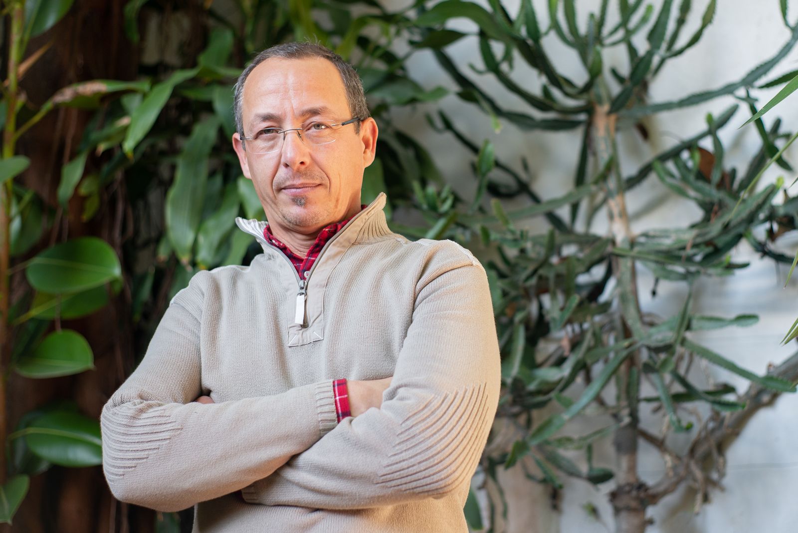 A confident man with eyeglasses posing outdoors with arms crossed, surrounded by lush greenery.