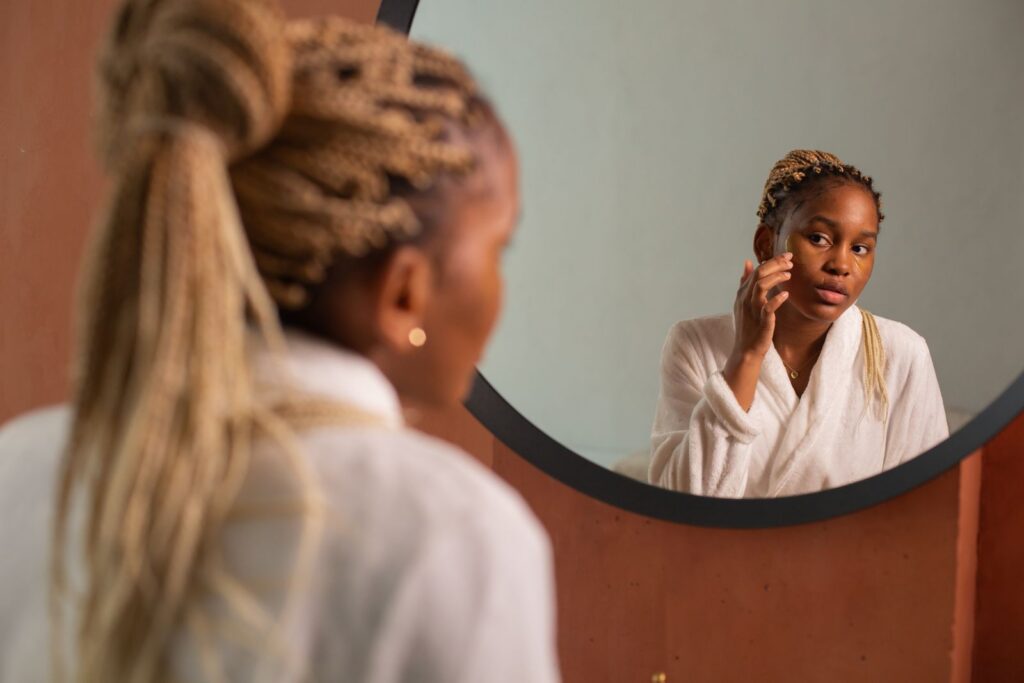 A woman with braided hair examines her skin in a round mirror indoors.