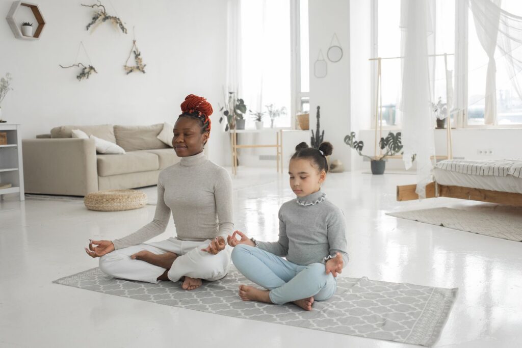 Full body of young African American woman with cute little daughter sitting in carpet with closed eyes and meditating in Lotus pose at home