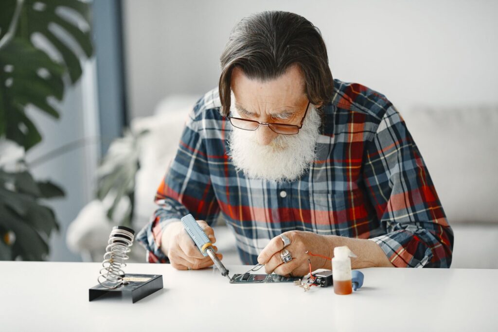Elderly man with beard focused on fixing electronic device indoors.