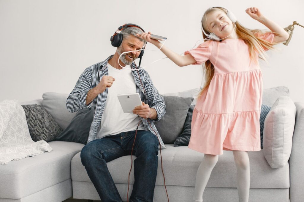 Father and daughter enjoying music while dancing on the couch with headphones in a living room.