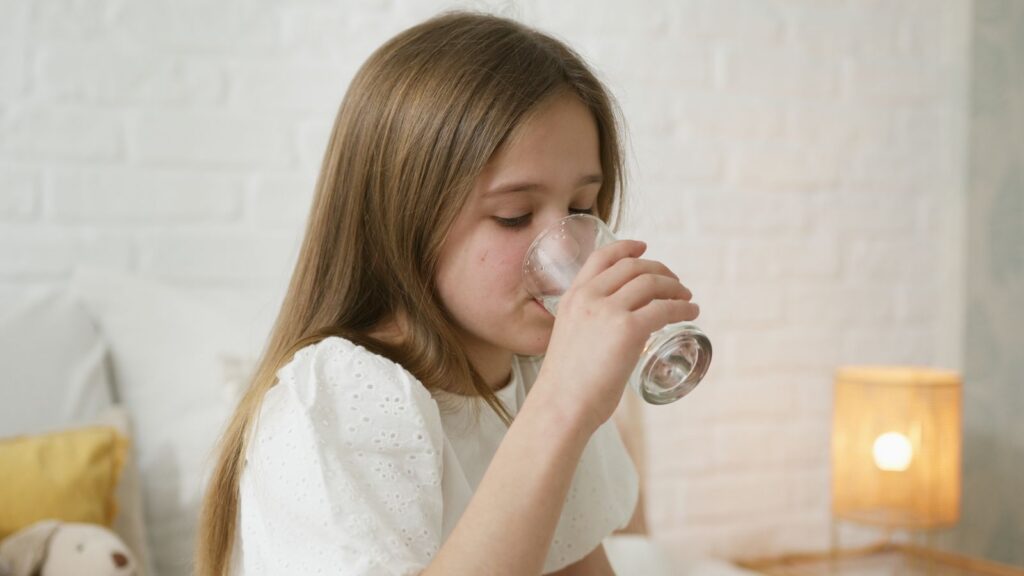 A young girl drinks water in a cozy bedroom, highlighting hydration and comfort.