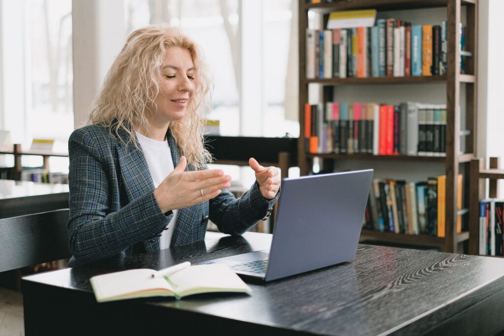 Positive adult female teacher with blond hair in elegant outfit sitting at table in library and explaining lesson during distant studies