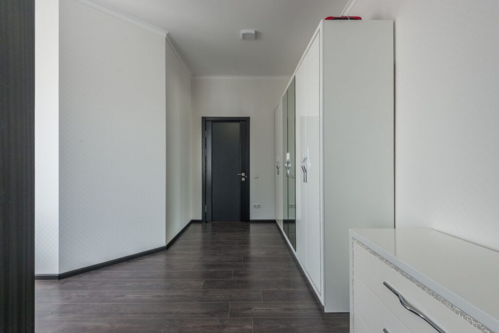 Interior of empty spacious hallway with wooden floor and white painted walls with black door on entrance and white wardrobe and commode