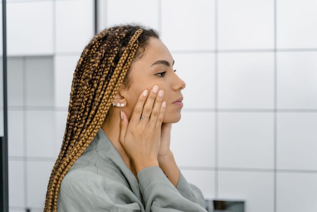 Profile view of a woman with braided hair gently touching her face, standing in front of a mirror.