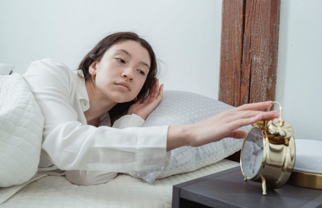 Sleepy young brunette female in white sleepwear with alarm clock not getting enough sleep