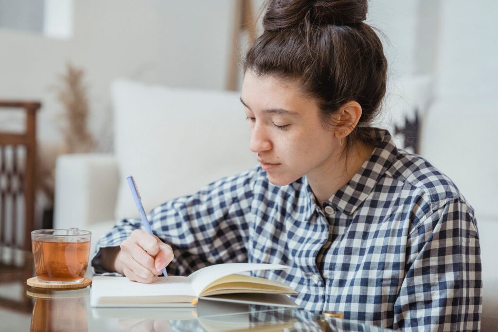 A young woman studying and writing in a notebook with a cup of tea nearby.