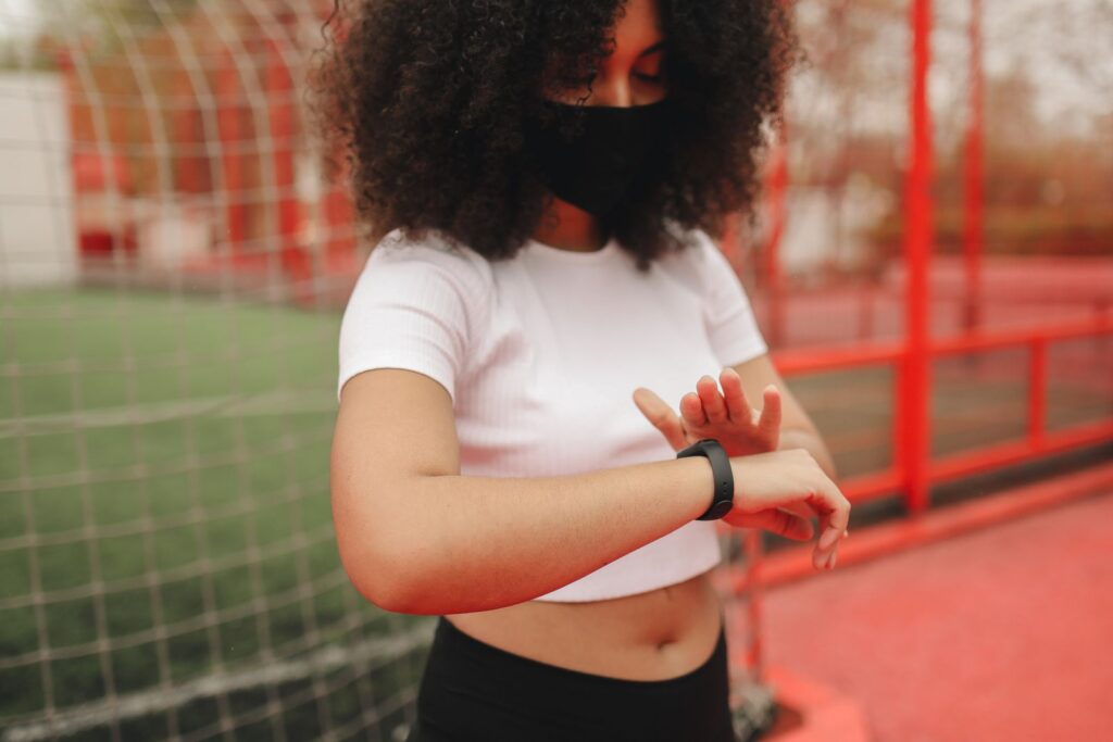 A woman with curly hair wearing a face mask checks the time on her smartwatch outside.
