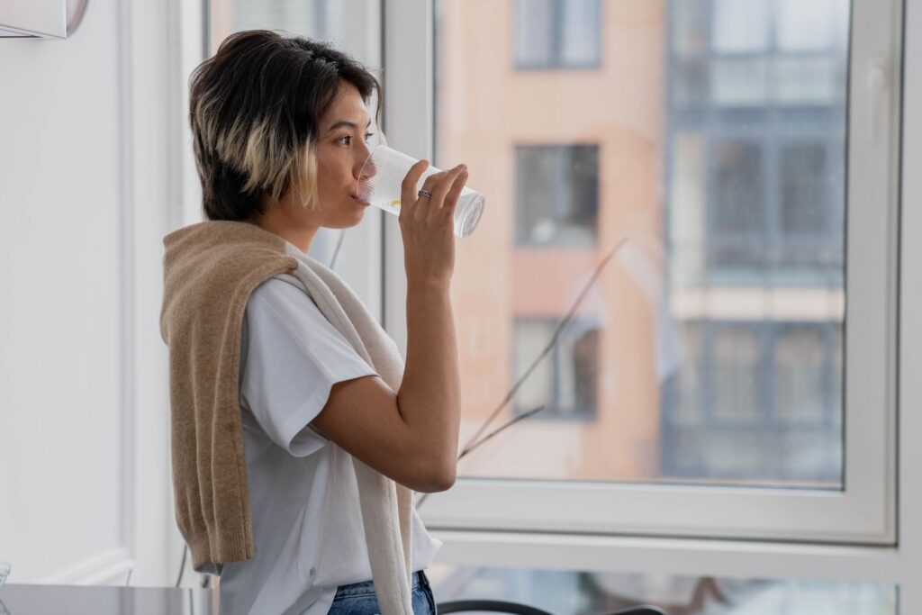 Asian woman drinking water near a window in a modern indoor setting, enjoying a refreshing moment.