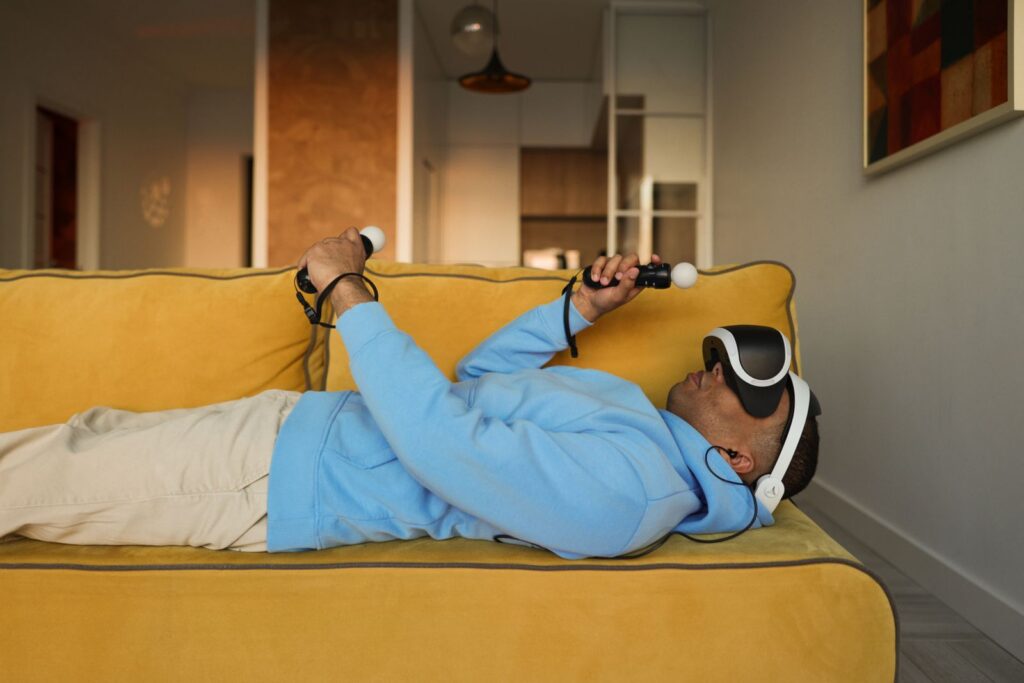 A man in a hoodie enjoys virtual reality gaming, lying on a yellow couch indoors.