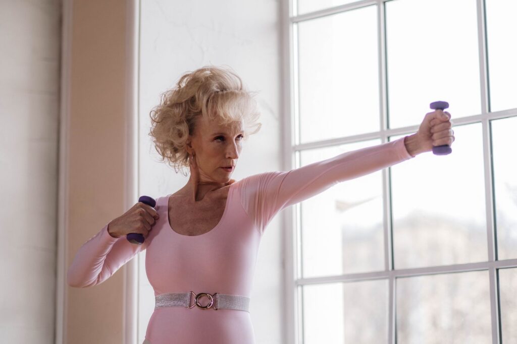 Senior woman in pink leotard working out with dumbbells near a large window indoors.