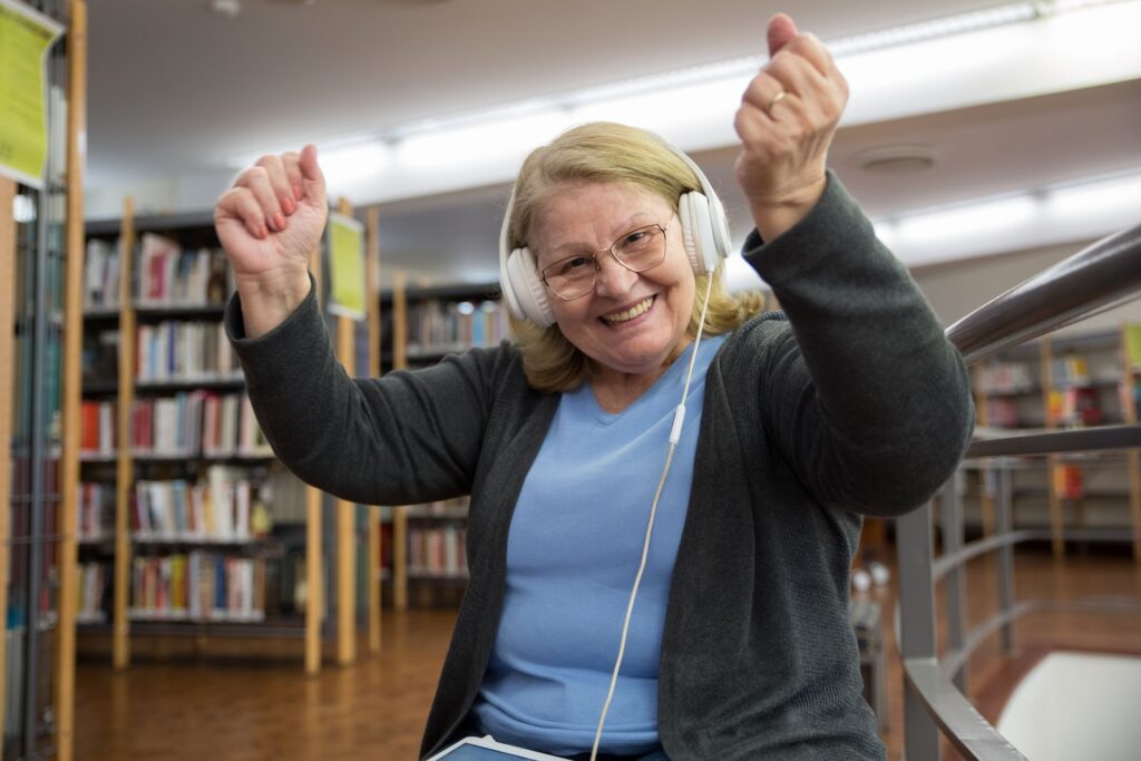 Smiling senior woman with headphones joyfully dancing in a cozy library setting.