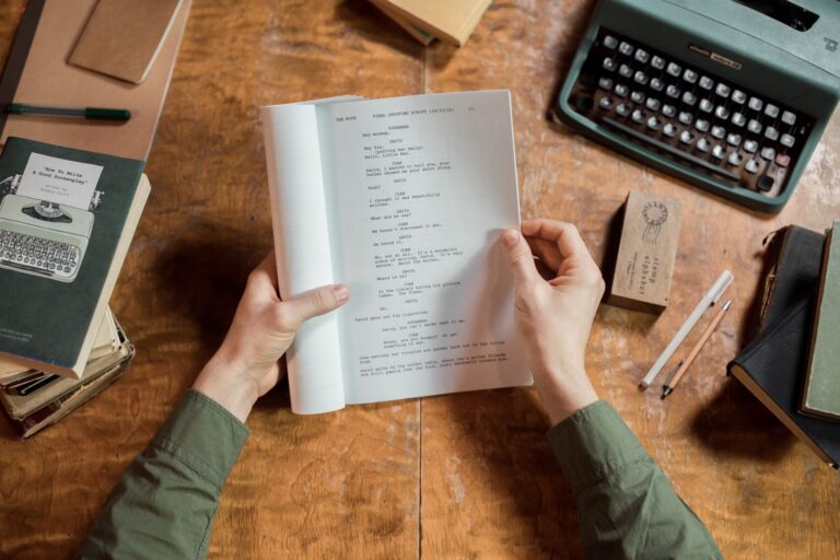 Hands holding script page at vintage desk with typewriter and books.