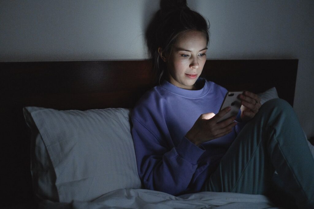A woman using her smartphone in bed at night, illuminated by the device's light.