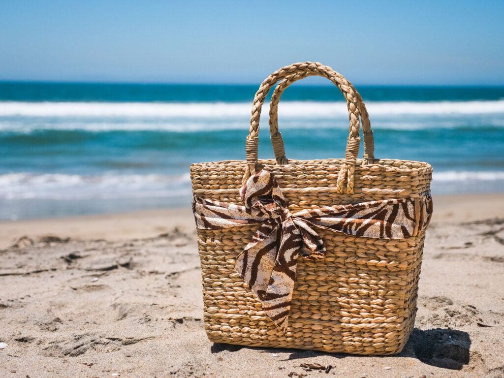 A stylish woven beach bag sits on a sunny beach with waves in the background.