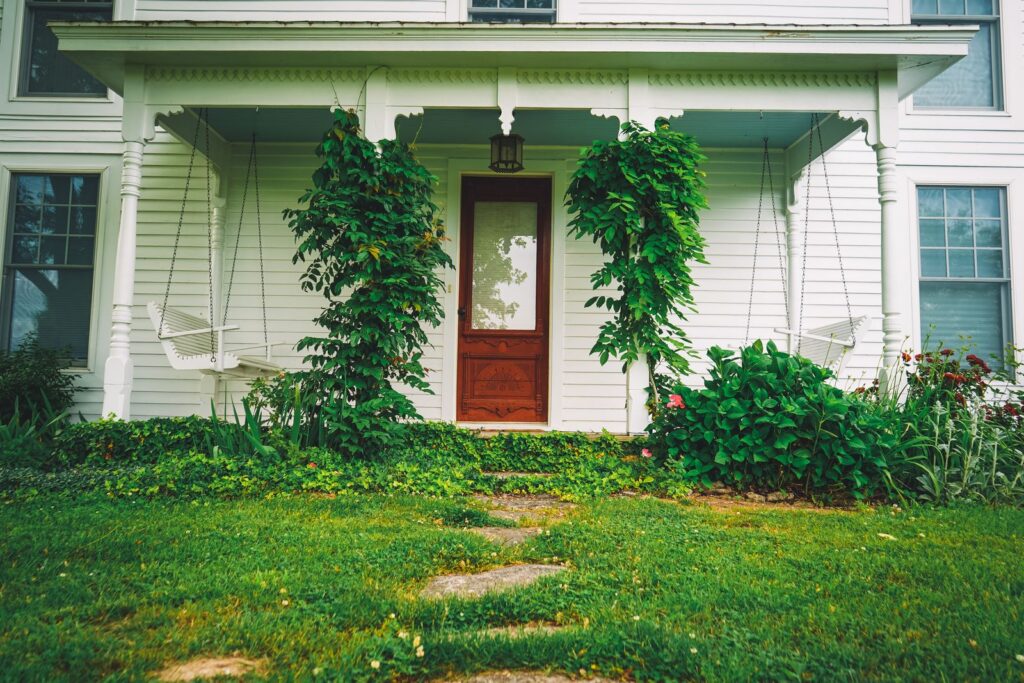 Inviting front porch of a white house with vibrant plants and two swings, showcasing a peaceful garden retreat.