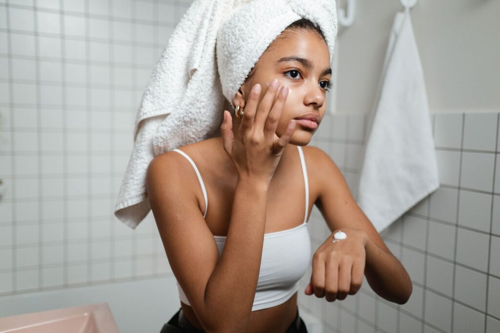 A young woman in a bathroom applying moisturizing cream with a towel on her head, focusing on self-care.