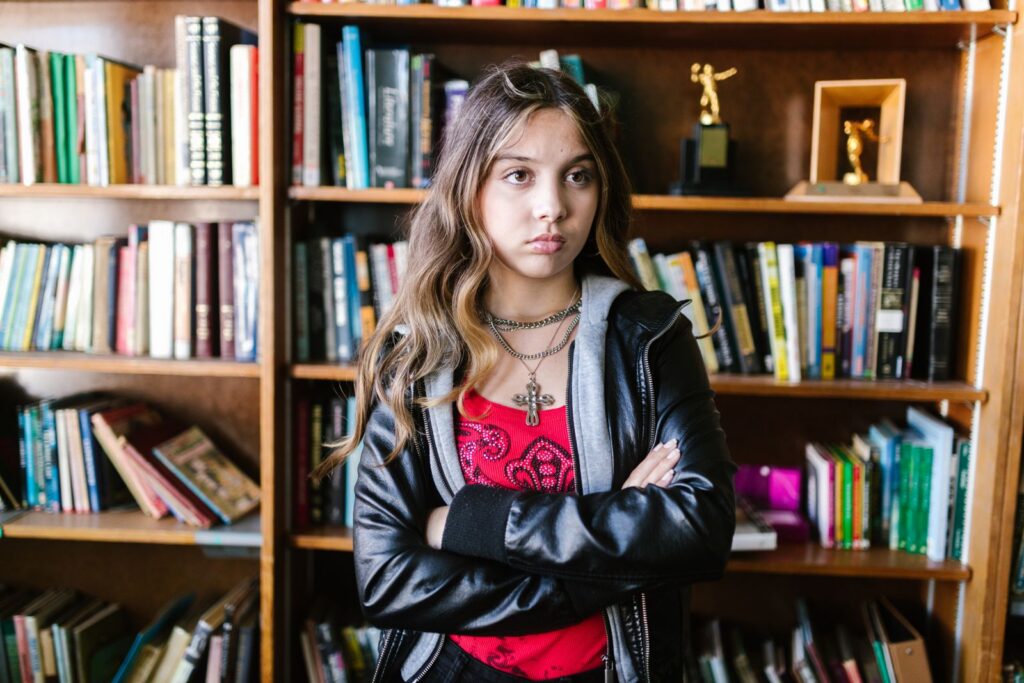 Teen girl with a leather jacket and arms crossed stands confidently in a library setting.