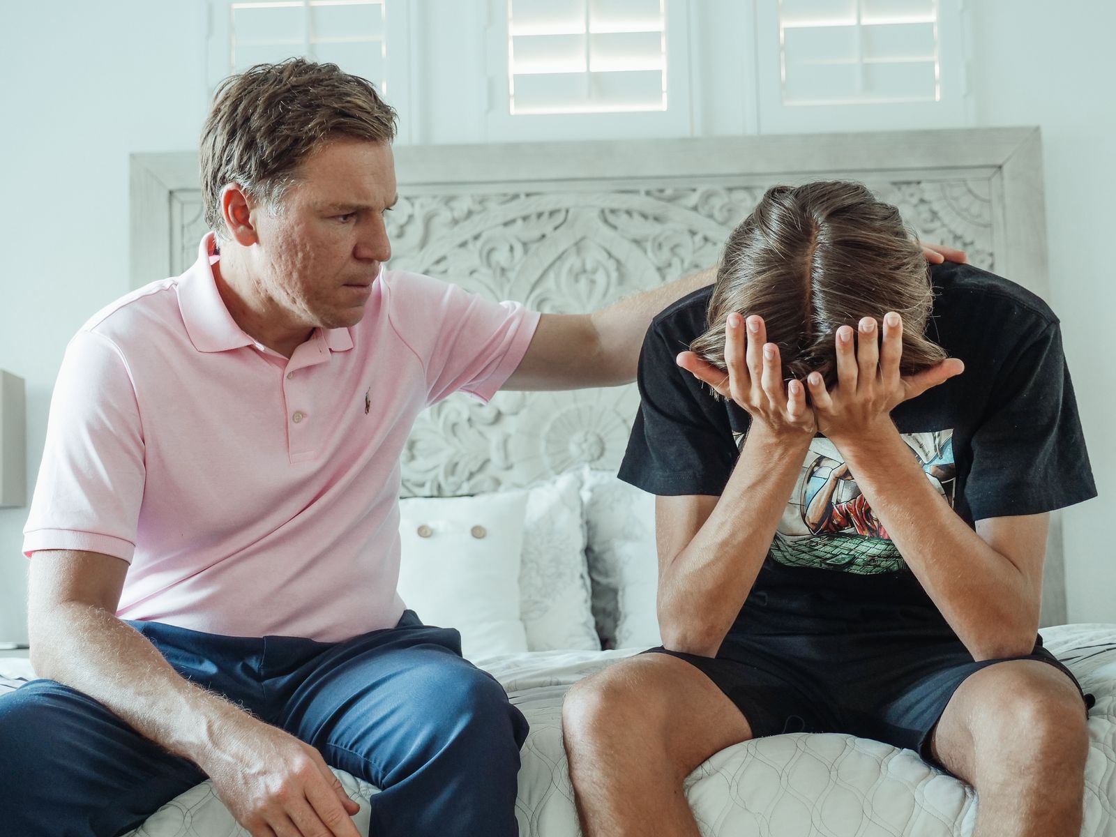 A compassionate father consoles his upset teenage son on a bed indoors.