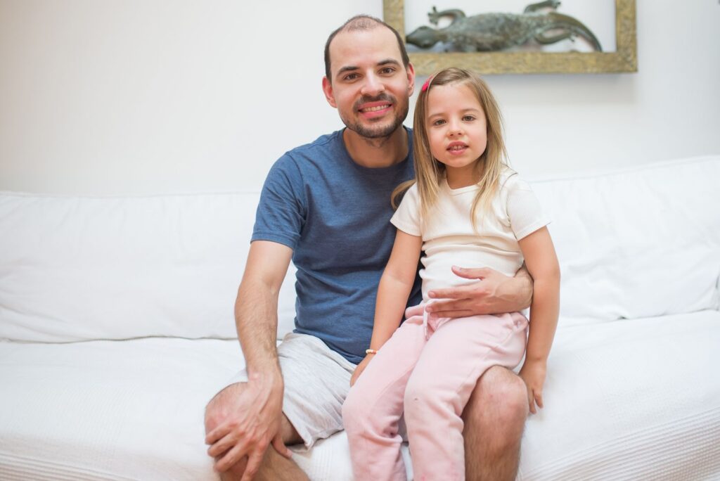 A father and daughter share a happy moment indoors, sitting on a sofa.