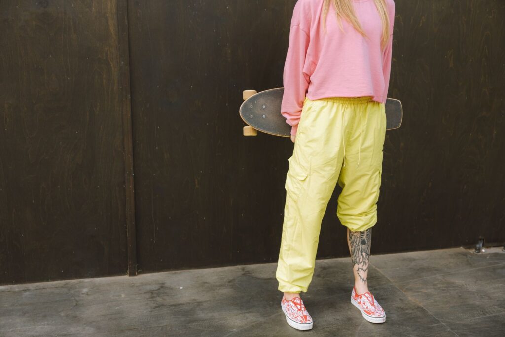 A young, tattooed skateboarder wearing a pink shirt and yellow pants holds a skateboard.