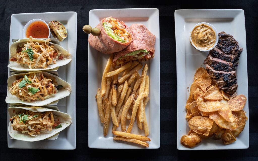 A top view of diverse dishes including tacos, a wrap with fries, and steak with chips on white plates.