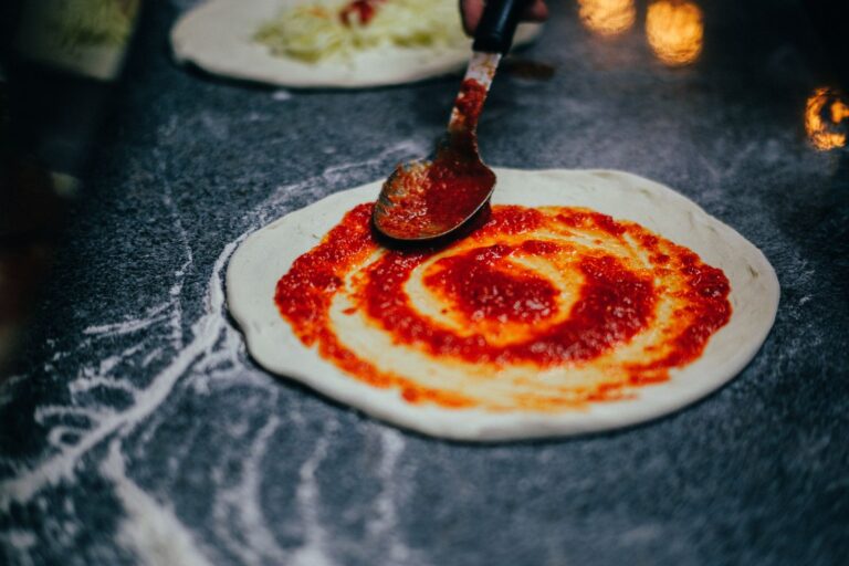 Preparing pizza dough with tomato sauce using a spoon in a kitchen setting.