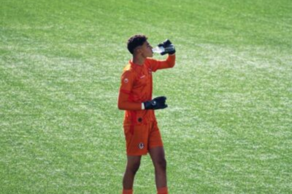 A soccer goalkeeper in orange attire takes a water break during a sunny day match on a green field.